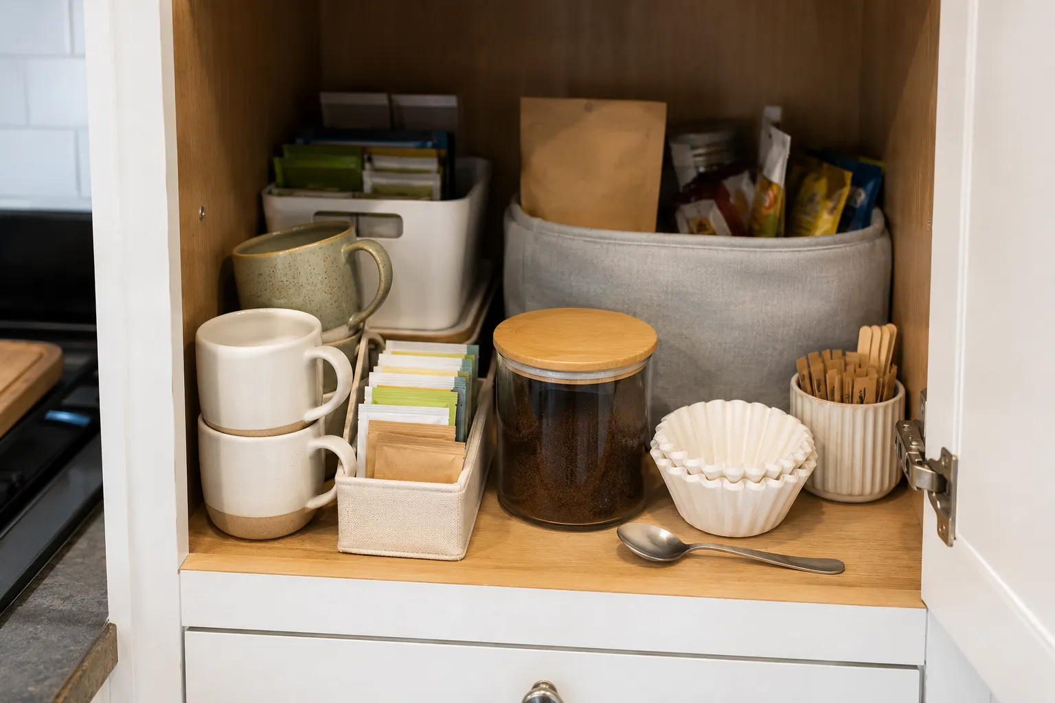 Small apartment kitchen cabinet shelf organized with mugs, tea bags, coffee supplies, filters, and simple storage bins.