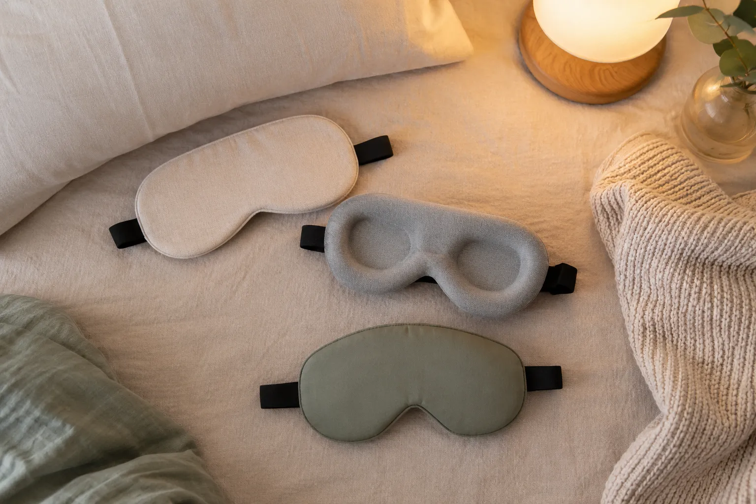 Flat lay of flat, contoured, and structured sleep masks on neutral bedding with soft bedroom lighting.